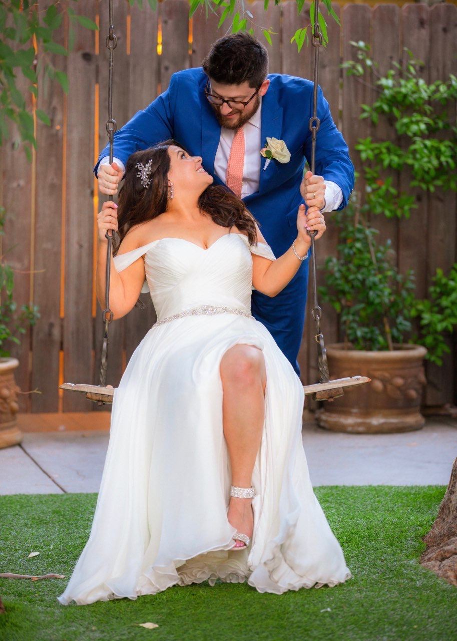 Bride In White Wedding Dress Siting On A Swing While Groom Pushes The Swing.