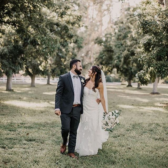 Bride In A White Wedding Dress Walking Through Field Of Trees With Groom.