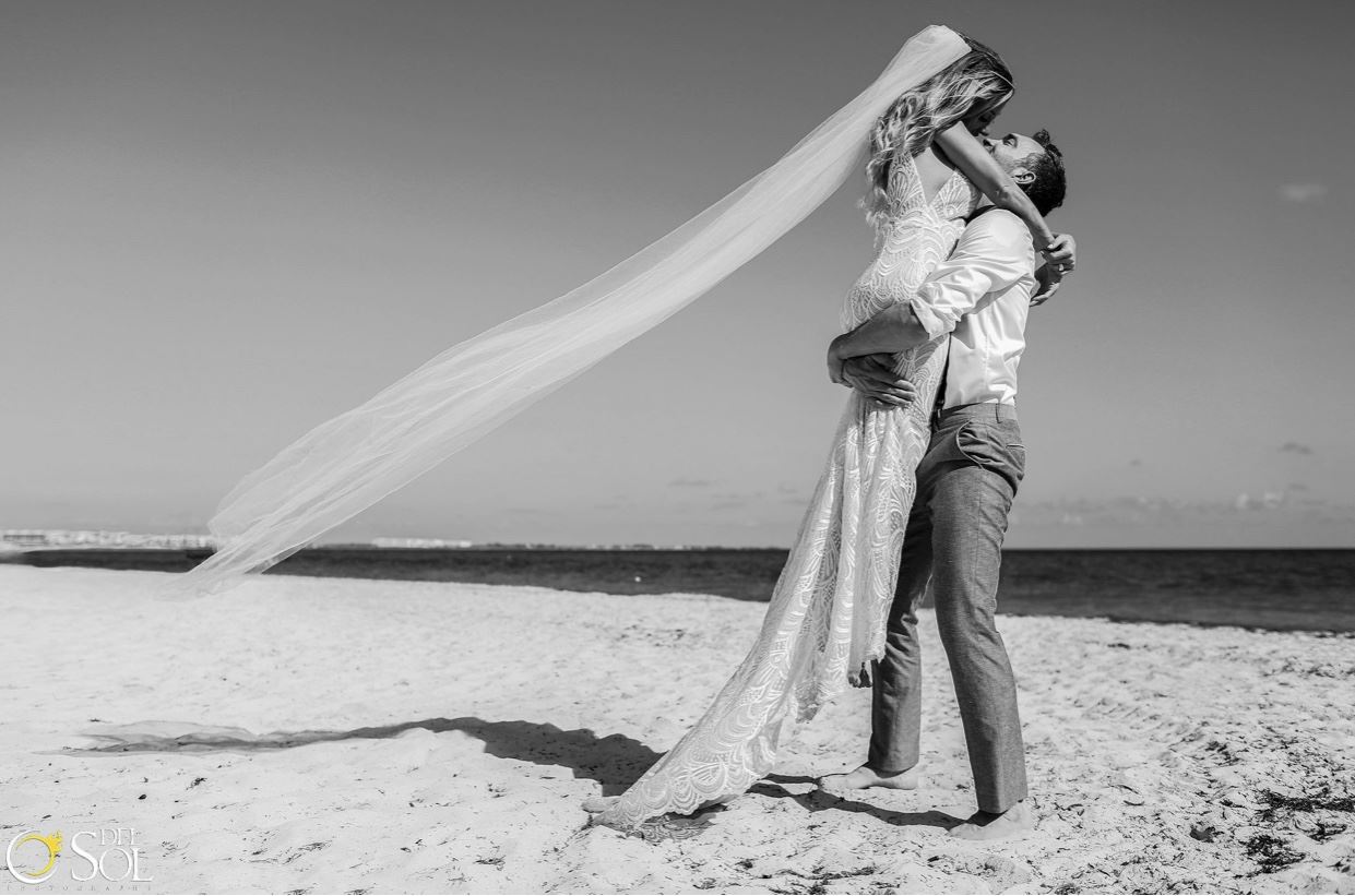 Groom Picking Up Bride On Sandy Beach. Veil Is Flowing In The Wind.