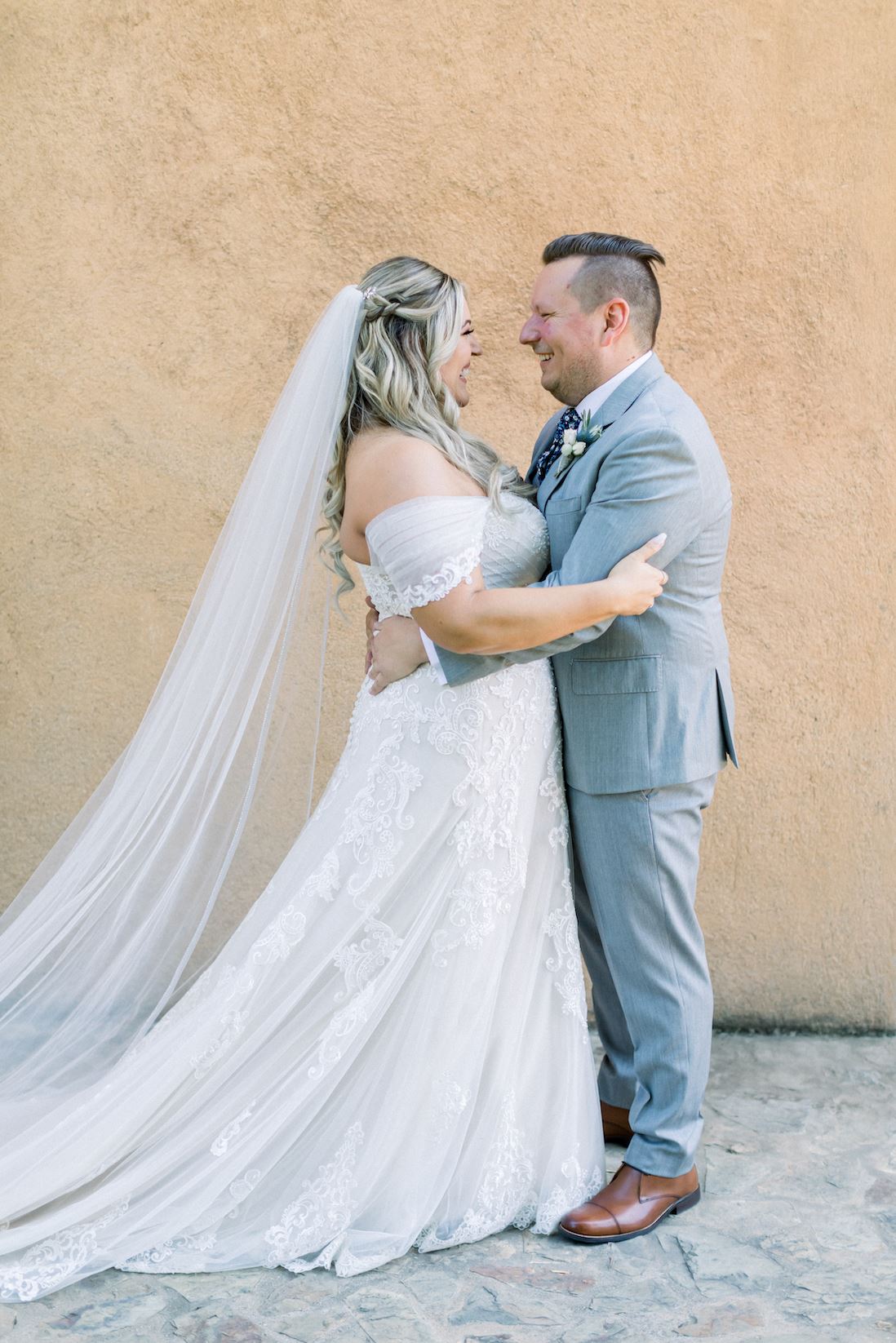 Bride In A White Wedding Dress Being Hugged By Groom In Grey Suit.
